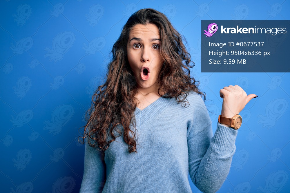 Young beautiful woman with curly hair wearing blue casual sweater over isolated background Surprised pointing with hand finger to the side, open mouth amazed expression.