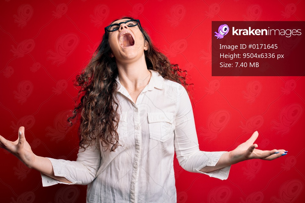 Young beautiful woman with curly hair wearing shirt and glasses over red background crazy and mad shouting and yelling with aggressive expression and arms raised. Frustration concept.
