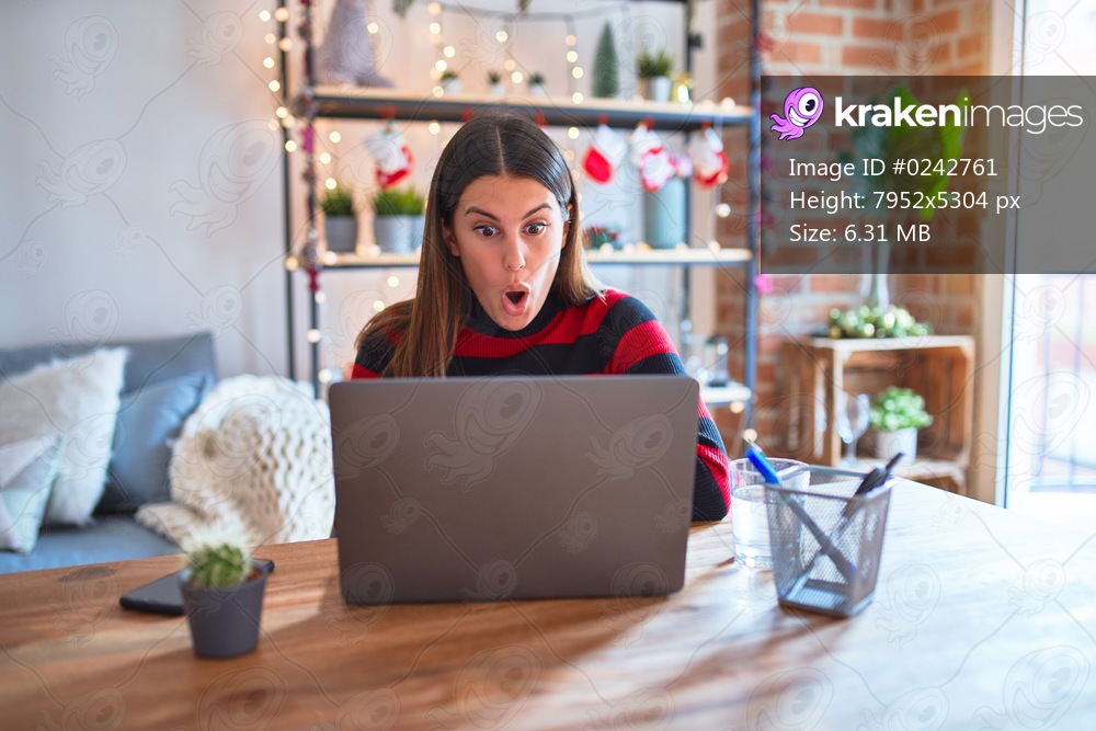 Beautiful woman sitting at the table working with laptop at home around christmas lights afraid and shocked with surprise expression, fear and excited face.