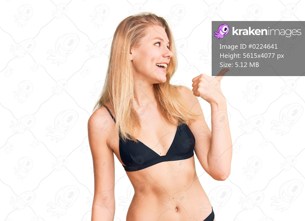 Young beautiful blonde woman wearing bikini smiling with happy face looking and pointing to the side with thumb up. 