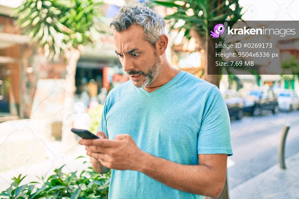 Middle age grey-haired man using smartphone at street of city scared and amazed with open mouth for surprise, disbelief face 