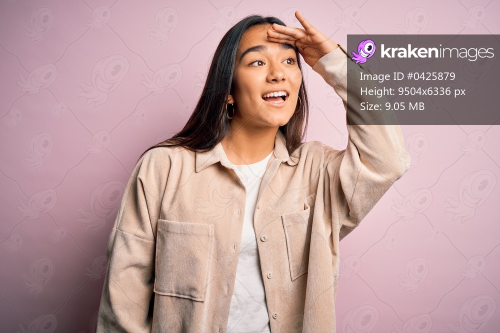 Young beautiful asian woman wearing casual shirt standing over pink background very happy and smiling looking far away with hand over head. Searching concept.