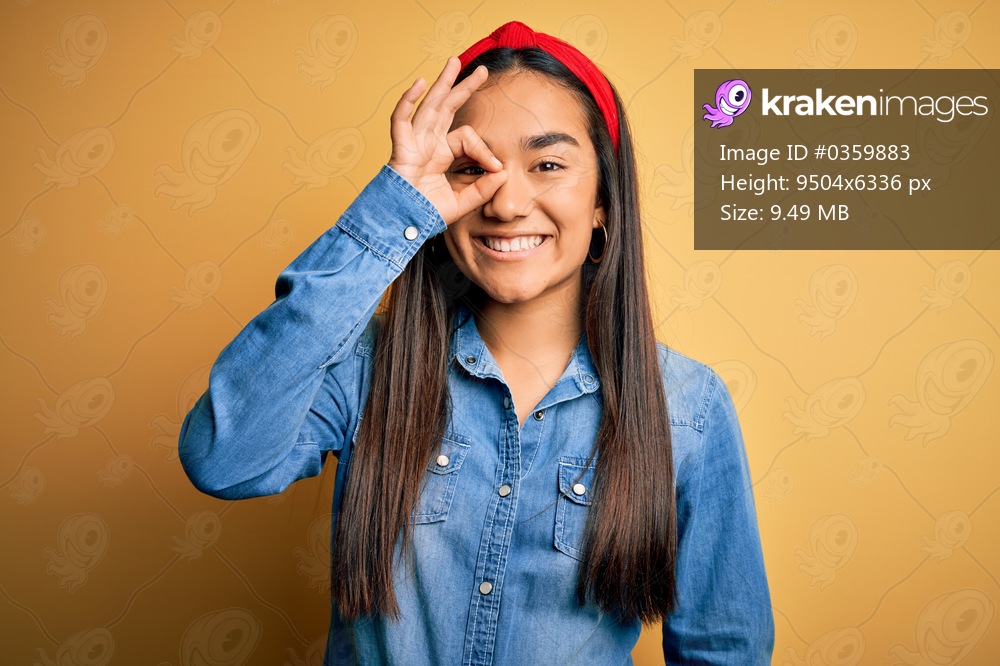 Young beautiful asian woman wearing casual denim shirt and diadem over yellow background doing ok gesture with hand smiling, eye looking through fingers with happy face.