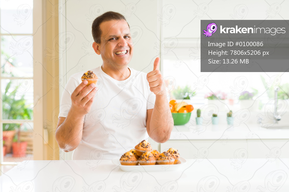 Middle age man eating chocolate chip muffins at home happy with big smile doing ok sign, thumb up with fingers, excellent sign