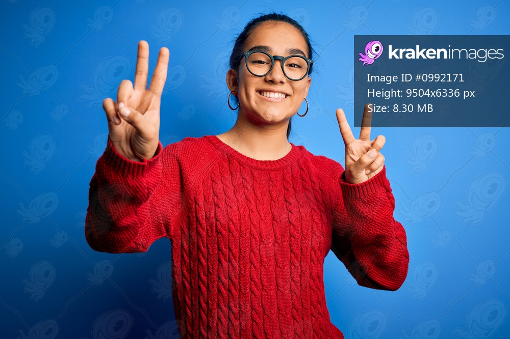Young beautiful asian woman wearing casual sweater and glasses over blue background smiling looking to the camera showing fingers doing victory sign. Number two.
