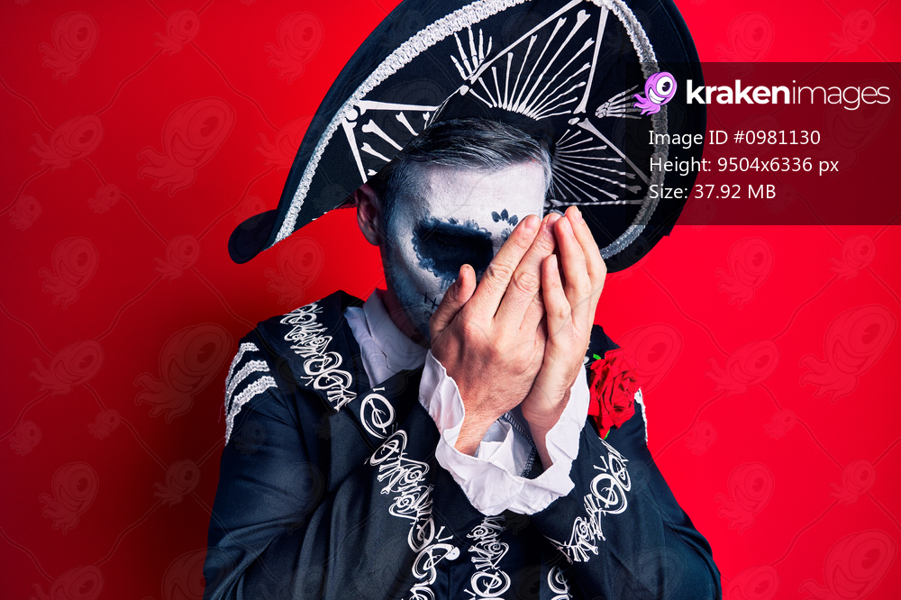 Young man wearing mexican day of the dead costume over red with sad expression covering face with hands while crying. depression concept. 