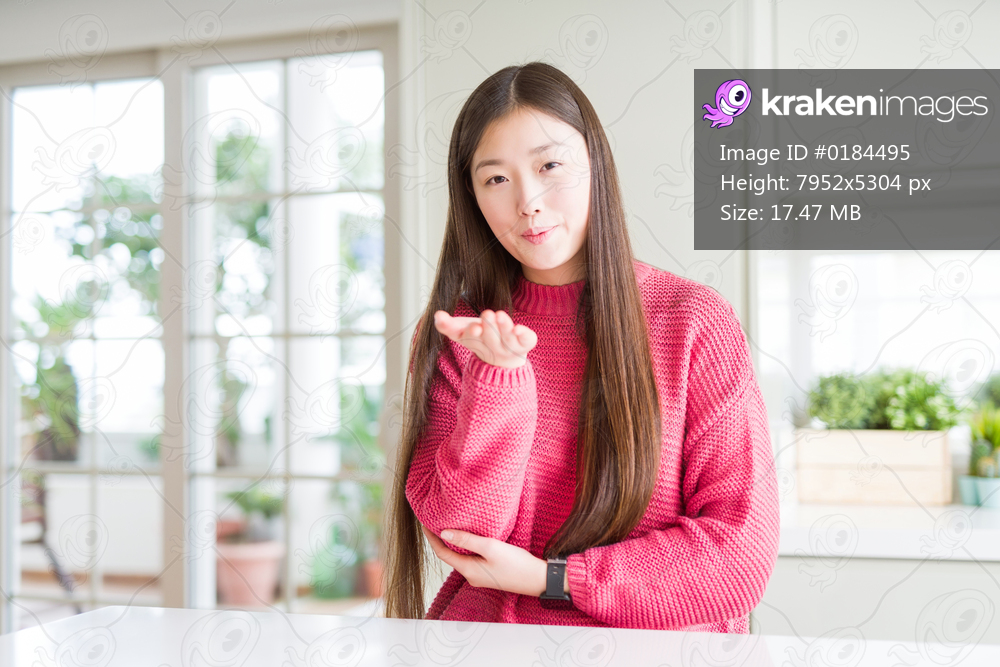 Beautiful Asian woman wearing pink sweater on white table looking at the camera blowing a kiss with hand on air being lovely and sexy. Love expression.