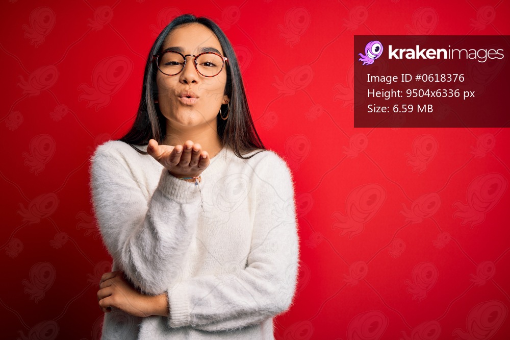 Young beautiful asian woman wearing casual sweater and glasses over red background looking at the camera blowing a kiss with hand on air being lovely and sexy. Love expression.