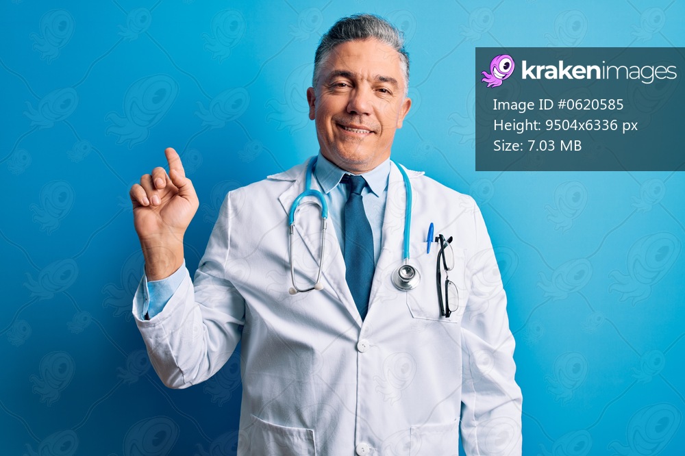 Middle age handsome grey-haired doctor man wearing coat and blue stethoscope with a big smile on face, pointing with hand finger to the side looking at the camera.