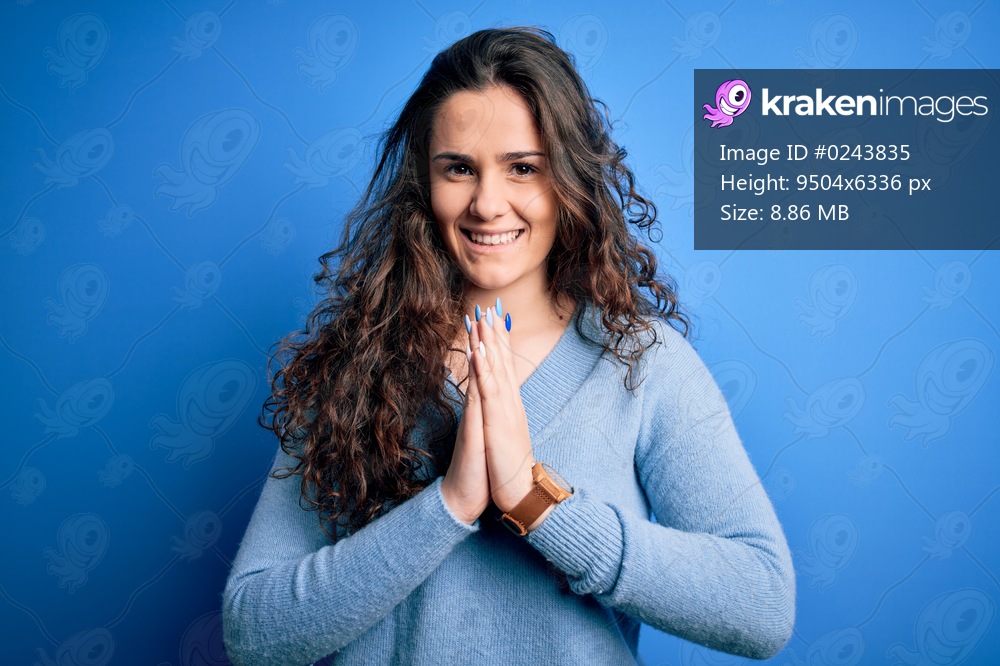 Young beautiful woman with curly hair wearing blue casual sweater over isolated background praying with hands together asking for forgiveness smiling confident.