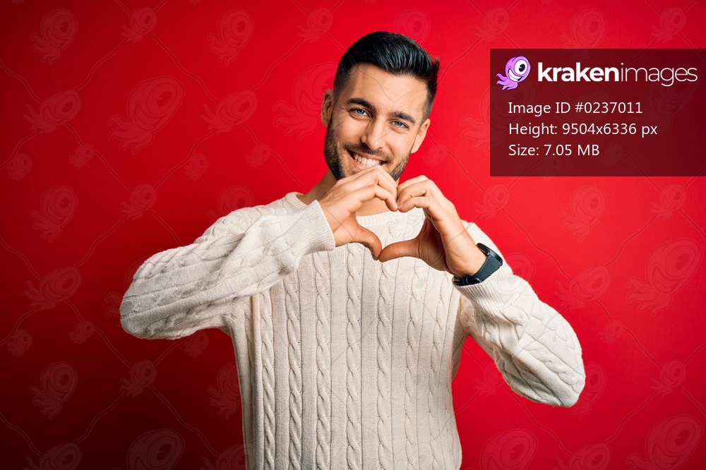 Young handsome man wearing casual white sweater standing over isolated red background smiling in love doing heart symbol shape with hands. Romantic concept.