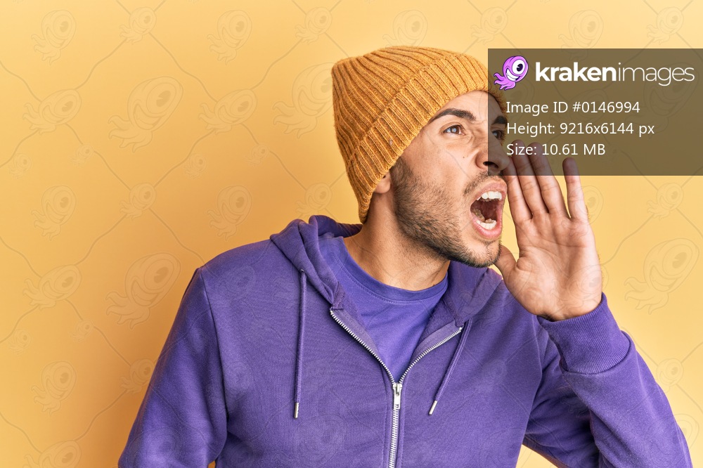 Young handsome man wearing wool winter hat shouting and screaming loud to side with hand on mouth. communication concept. 