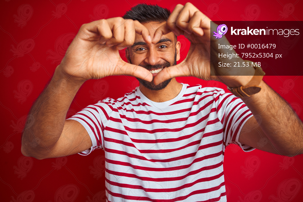 Young indian man wearing striped t-shirt standing over isolated red background smiling in love showing heart symbol and shape with hands. Romantic concept.