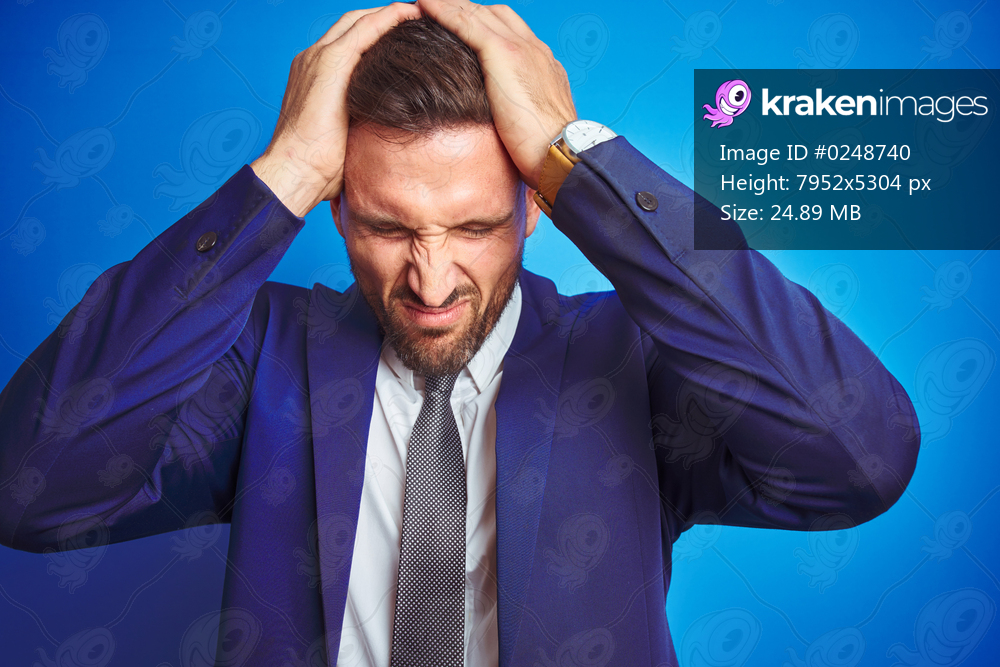 Close up picture of young handsome business man over blue isolated background suffering from headache desperate and stressed because pain and migraine. Hands on head.