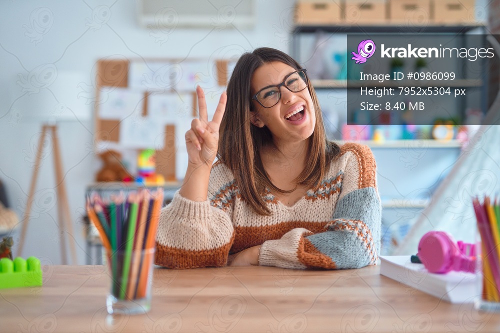 Young beautiful teacher woman wearing sweater and glasses sitting on desk at kindergarten smiling looking to the camera showing fingers doing victory sign. Number two.