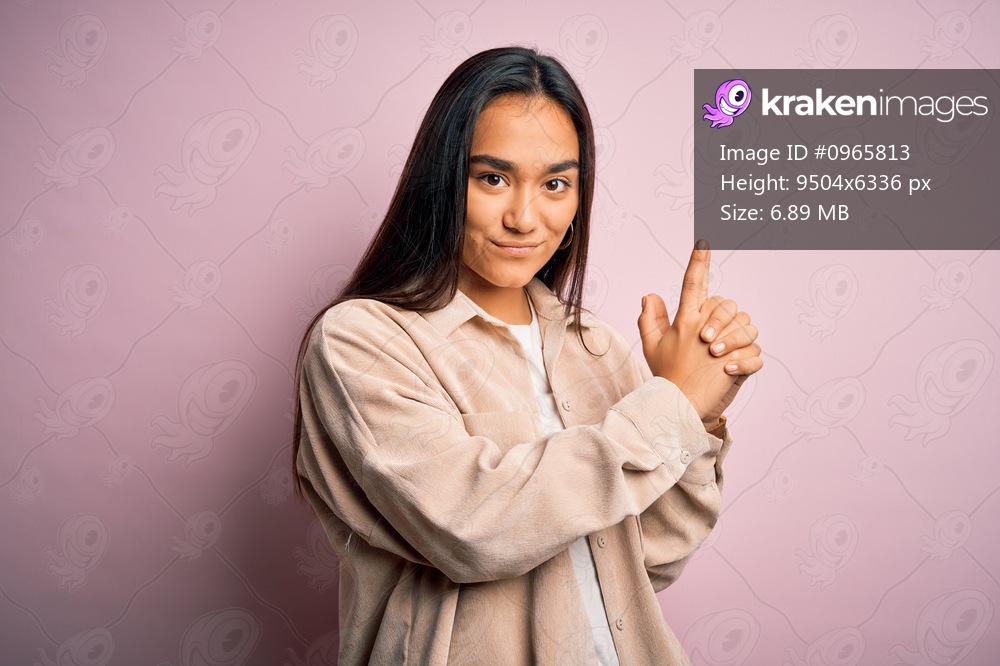 Young beautiful asian woman wearing casual shirt standing over pink background Holding symbolic gun with hand gesture, playing killing shooting weapons, angry face