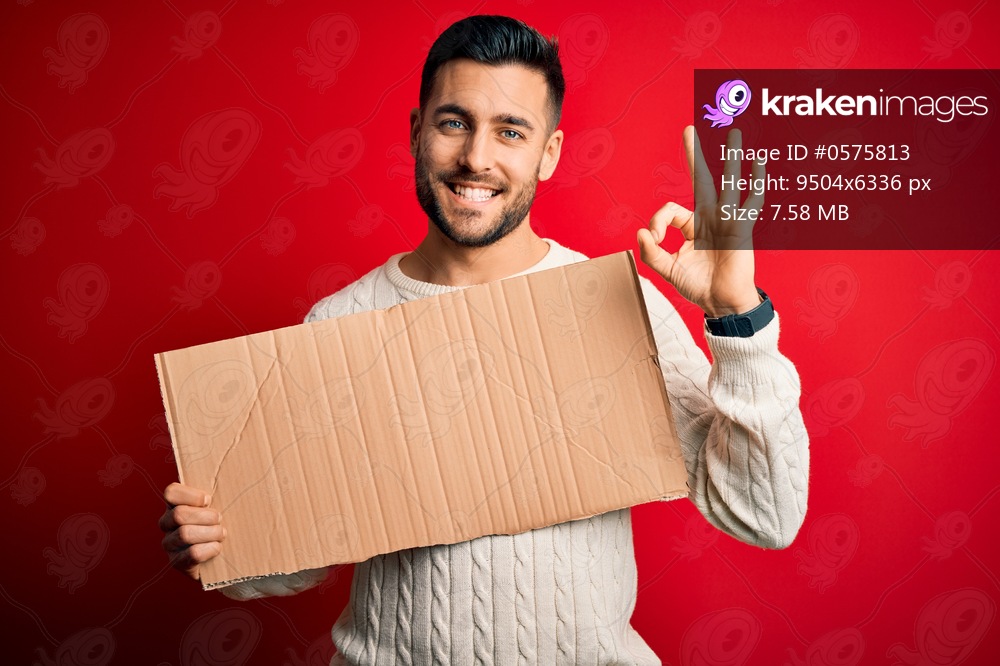 Young handsome man holding blank cardboard banner over isolated red background doing ok sign with fingers, excellent symbol