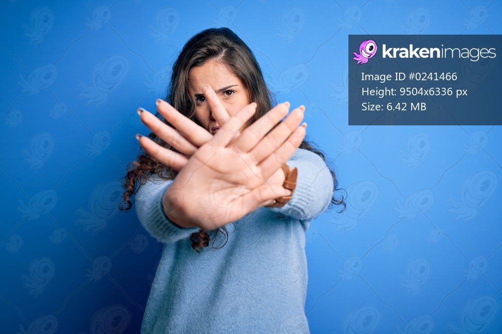 Young beautiful woman with curly hair wearing blue casual sweater over isolated background Rejection expression crossing arms and palms doing negative sign, angry face