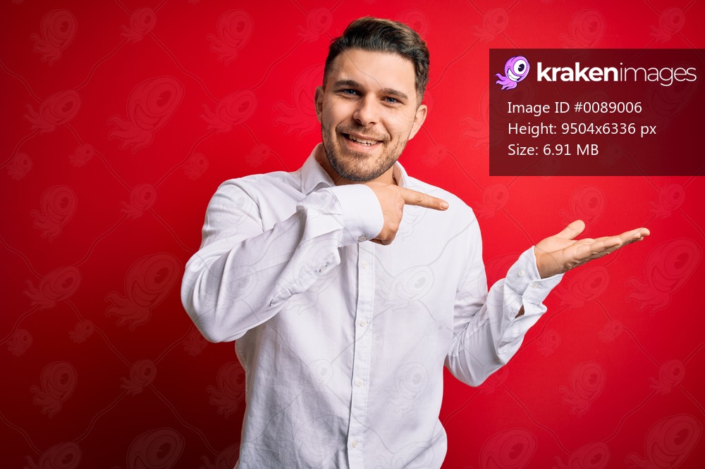Young business man with blue eyes wearing elegant shirt standing over red isolated background amazed and smiling to the camera while presenting with hand and pointing with finger.