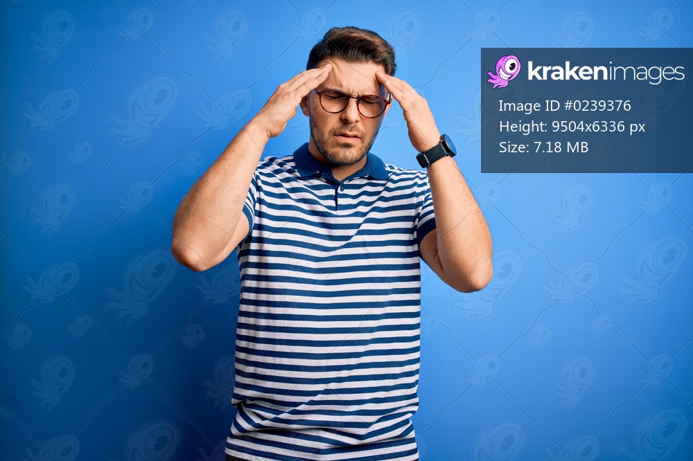 Young man with blue eyes wearing glasses and casual striped t-shirt over blue background with hand on head for pain in head because stress. Suffering migraine.