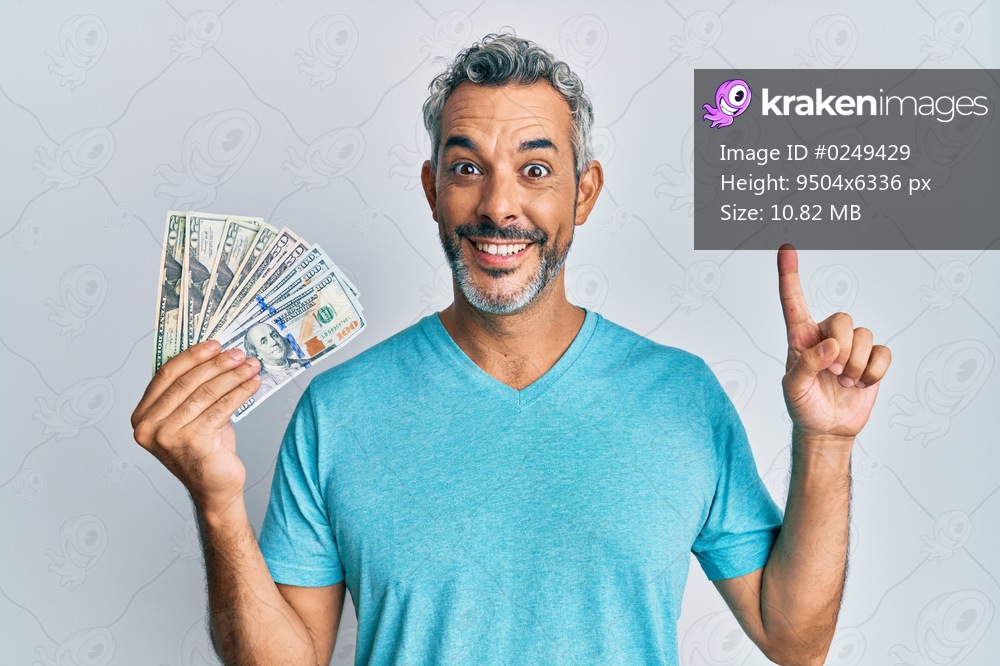 Middle age grey-haired man holding dollars smiling with an idea or question pointing finger with happy face, number one 