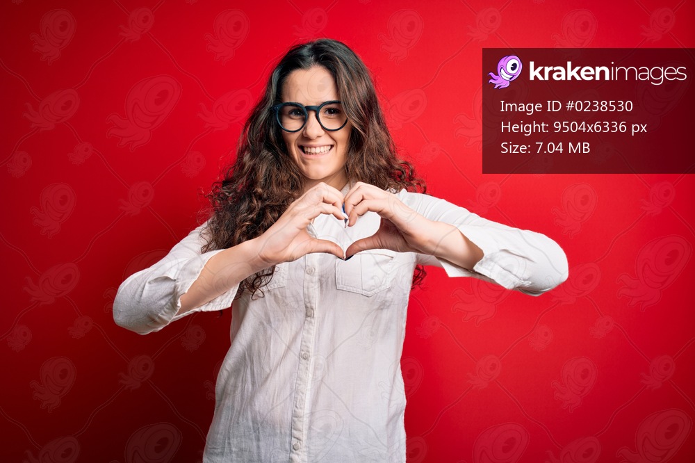 Young beautiful woman with curly hair wearing shirt and glasses over red background smiling in love showing heart symbol and shape with hands. Romantic concept.