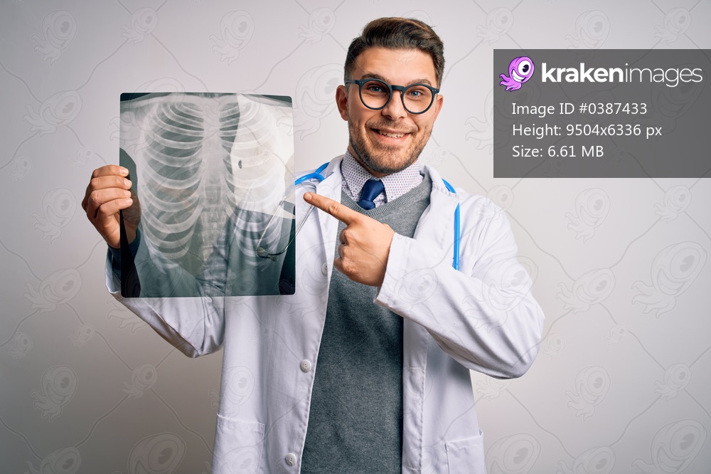 Young doctor man wearing medical coat looking at chest scan radiography over isolated background very happy pointing with hand and finger