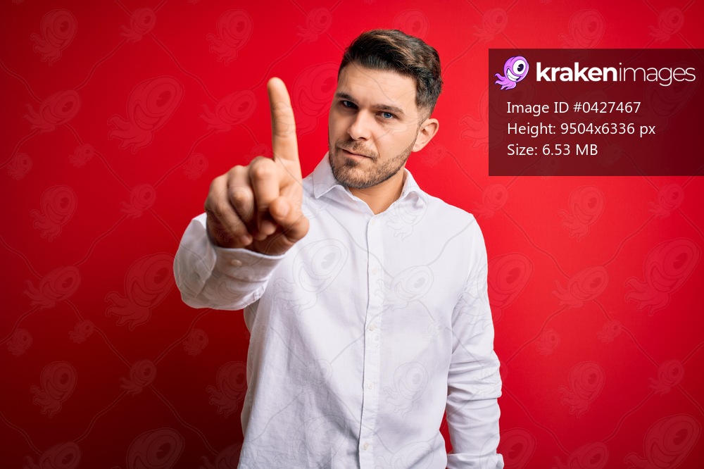 Young business man with blue eyes wearing elegant shirt standing over red isolated background Pointing with finger up and angry expression, showing no gesture