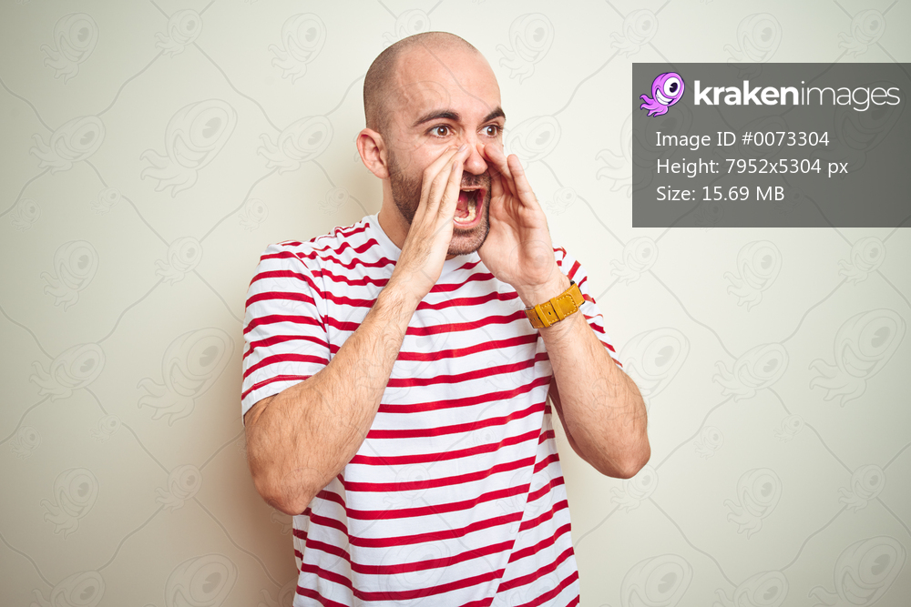 Young bald man with beard wearing casual striped red t-shirt over white isolated background Shouting angry out loud with hands over mouth