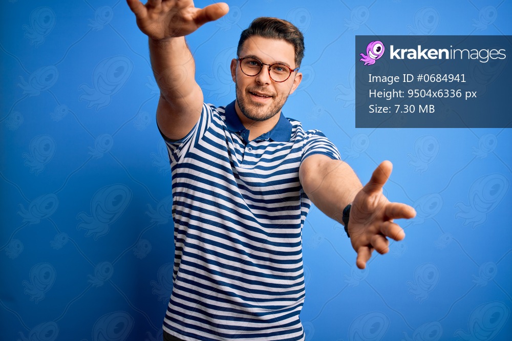 Young man with blue eyes wearing glasses and casual striped t-shirt over blue background looking at the camera smiling with open arms for hug. Cheerful expression embracing happiness.