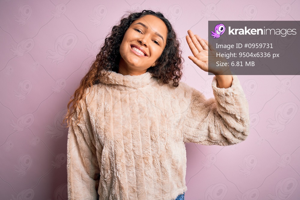 Young beautiful woman with curly hair wearing casual sweater standing over pink background Waiving saying hello happy and smiling, friendly welcome gesture