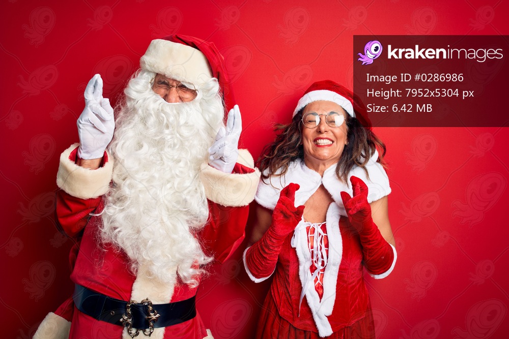 Middle age couple wearing Santa costume and glasses over isolated red background gesturing finger crossed smiling with hope and eyes closed. Luck and superstitious concept.
