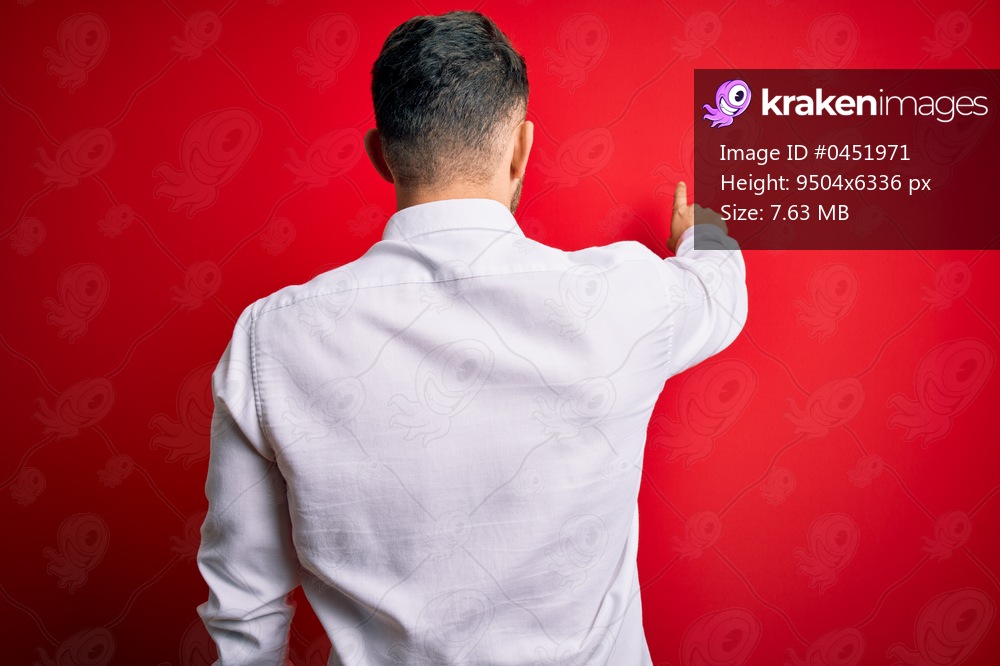 Young business man with blue eyes wearing elegant shirt standing over red isolated background Posing backwards pointing ahead with finger hand