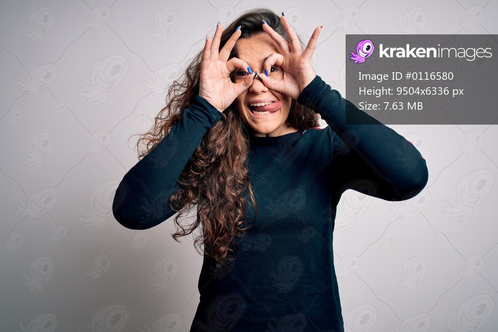 Young beautiful woman with curly hair wearing casual sweater over isolated white background doing ok gesture like binoculars sticking tongue out, eyes looking through fingers. Crazy expression.