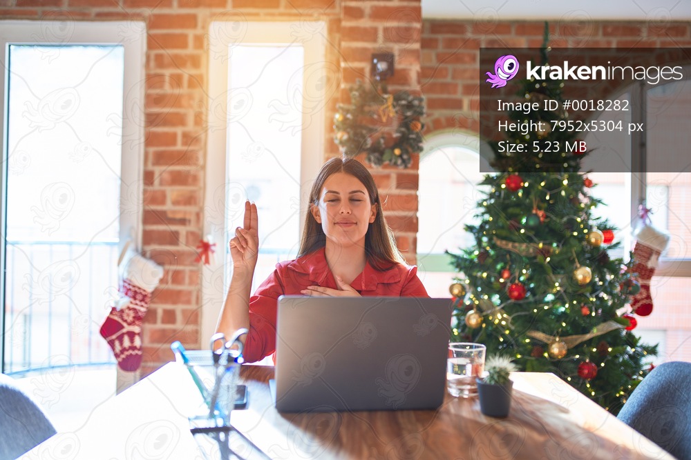 Beautiful woman sitting at the table working with laptop at home around christmas tree smiling swearing with hand on chest and fingers up, making a loyalty promise oath