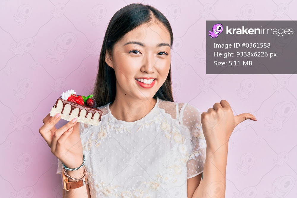 Young chinese woman eating a tasty cake pointing thumb up to the side smiling happy with open mouth 