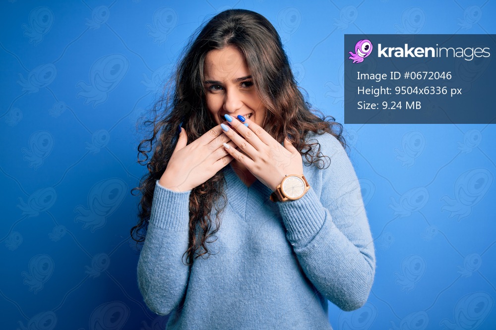 Young beautiful woman with curly hair wearing blue casual sweater over isolated background laughing and embarrassed giggle covering mouth with hands, gossip and scandal concept