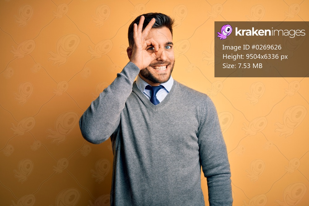 Young handsome businessman wearing elegant sweater and tie over yellow background doing ok gesture with hand smiling, eye looking through fingers with happy face.