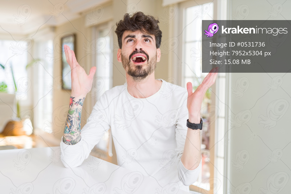 Young man wearing casual shirt sitting on white table crazy and mad shouting and yelling with aggressive expression and arms raised. Frustration concept.