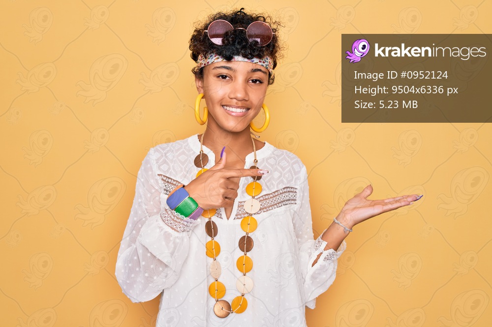 Young beautiful african american afro hippie woman wearing sunglasses and accessories amazed and smiling to the camera while presenting with hand and pointing with finger.