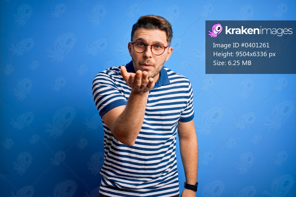 Young man with blue eyes wearing glasses and casual striped t-shirt over blue background looking at the camera blowing a kiss with hand on air being lovely and sexy. Love expression.
