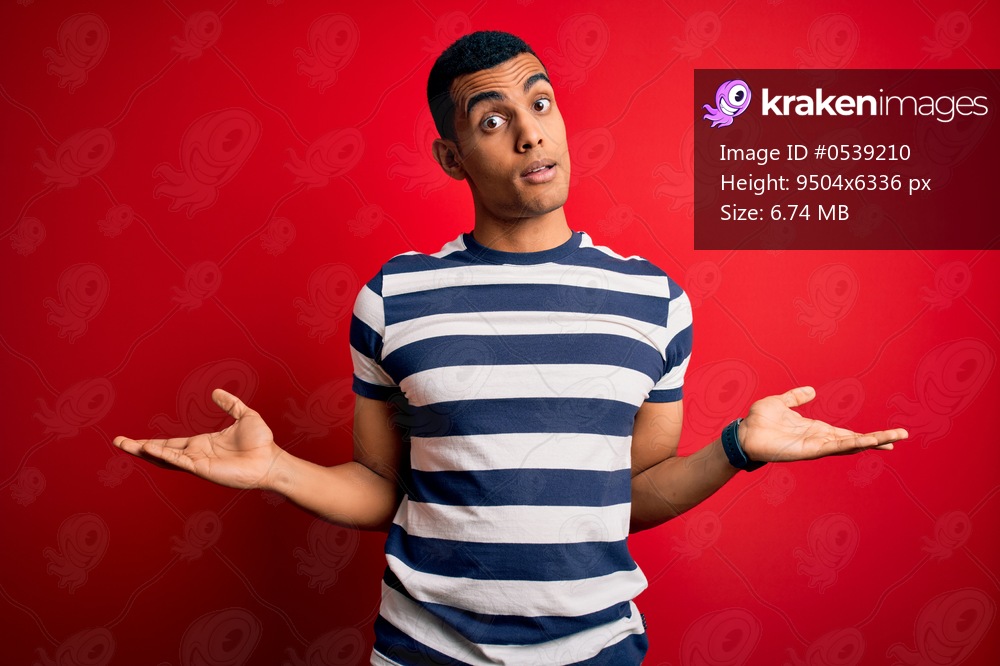 Handsome african american man wearing casual striped t-shirt standing over red background clueless and confused with open arms, no idea concept.