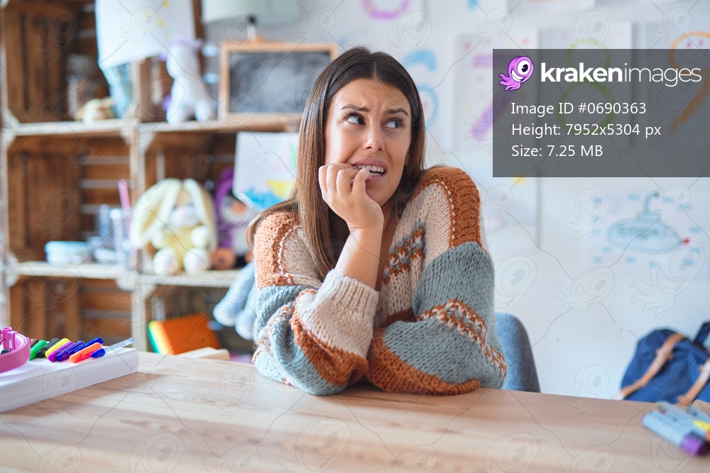 Young beautiful teacher woman wearing sweater and glasses sitting on desk at kindergarten looking stressed and nervous with hands on mouth biting nails. Anxiety problem.
