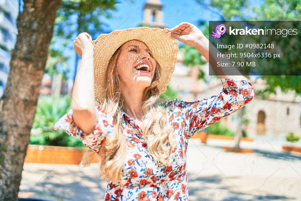 Young beautiful blonde woman on vacation wearing summer hat smiling happy. Standing with smile on face at street of city.