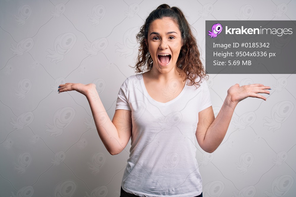 Young beautiful woman with curly hair wearing casual t-shirt standing over white background celebrating crazy and amazed for success with arms raised and open eyes screaming excited. Winner concept