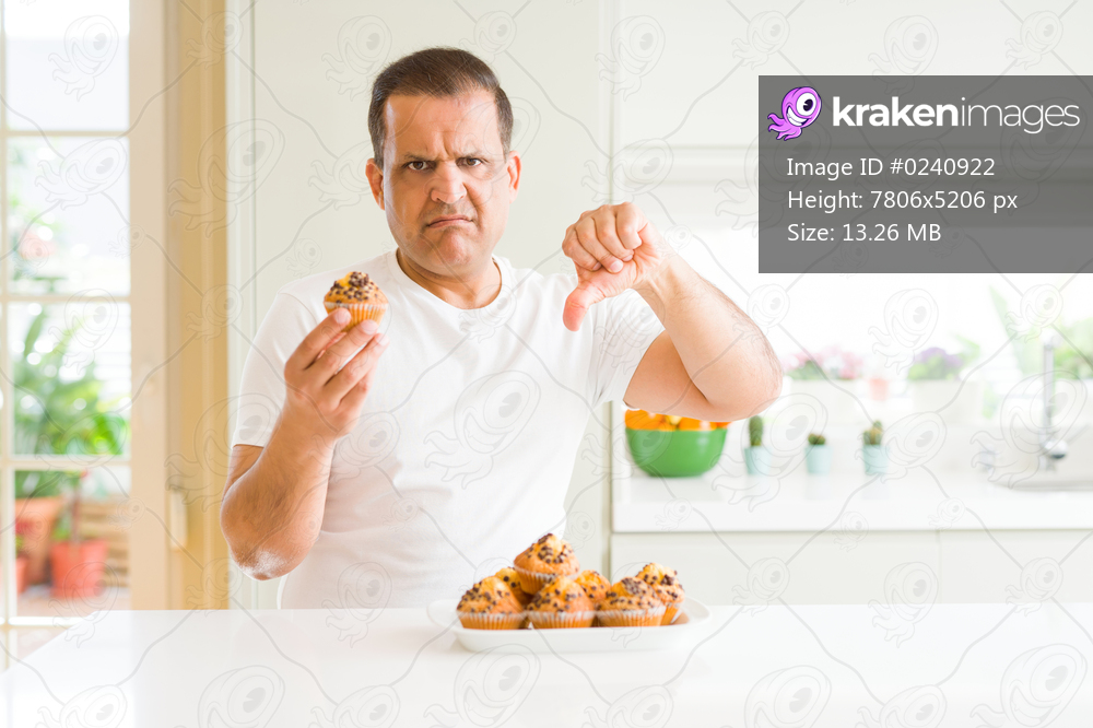 Middle age man eating chocolate chip muffins at home with angry face, negative sign showing dislike with thumbs down, rejection concept