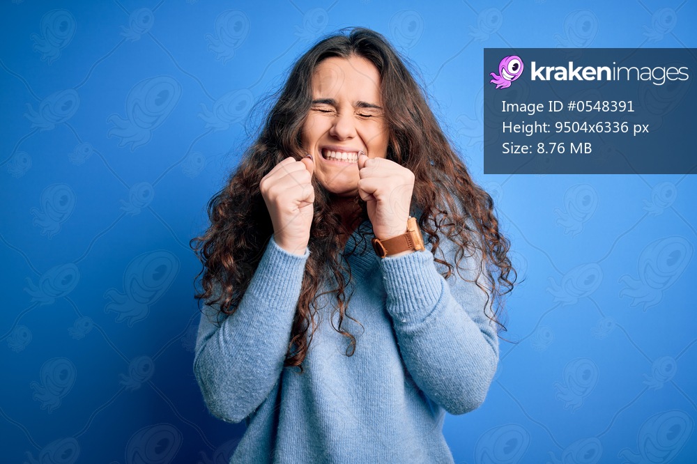 Young beautiful woman with curly hair wearing blue casual sweater over isolated background excited for success with arms raised and eyes closed celebrating victory smiling. Winner concept.