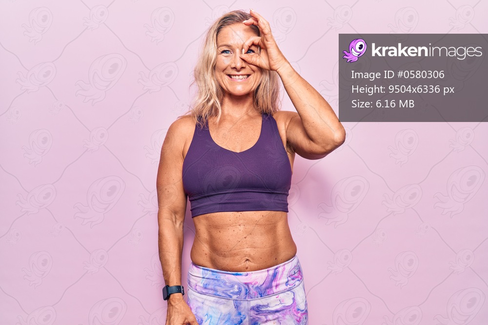 Middle age caucasian blonde woman wearing sportswear over pink background doing ok gesture with hand smiling, eye looking through fingers with happy face. 