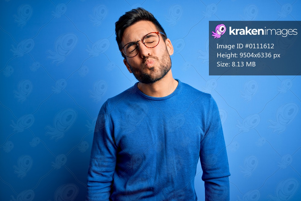 Young handsome man with beard wearing casual sweater and glasses over blue background looking at the camera blowing a kiss on air being lovely and sexy. Love expression.
