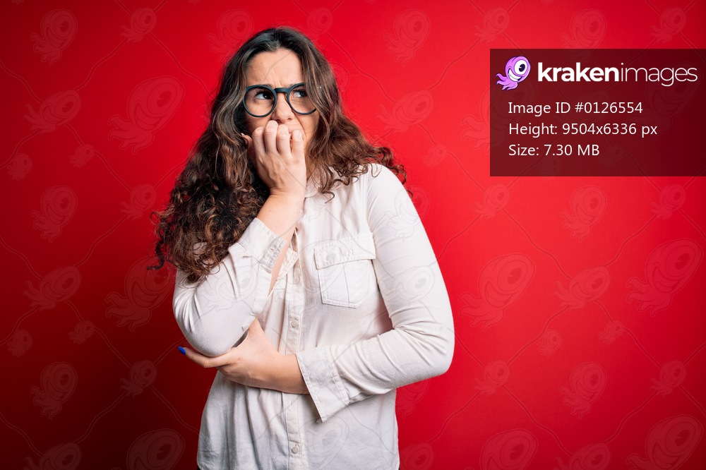 Young beautiful woman with curly hair wearing shirt and glasses over red background looking stressed and nervous with hands on mouth biting nails. Anxiety problem.